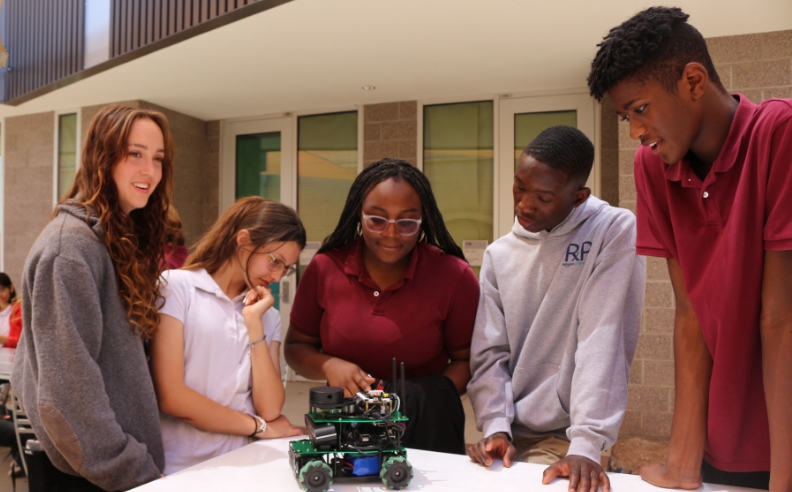 Five Fulton Schools students standing at a desk, observing a robot with wheels at the Polytechnic Robotics Demo event.