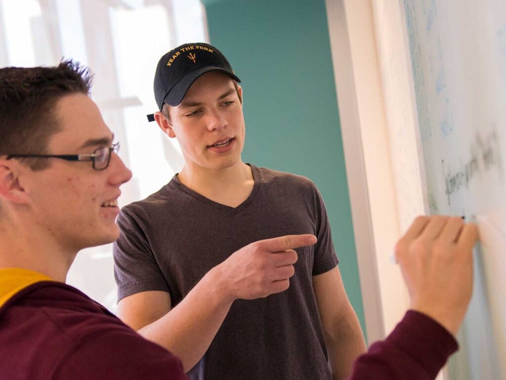 A student helps another student while they both stand at a white board.