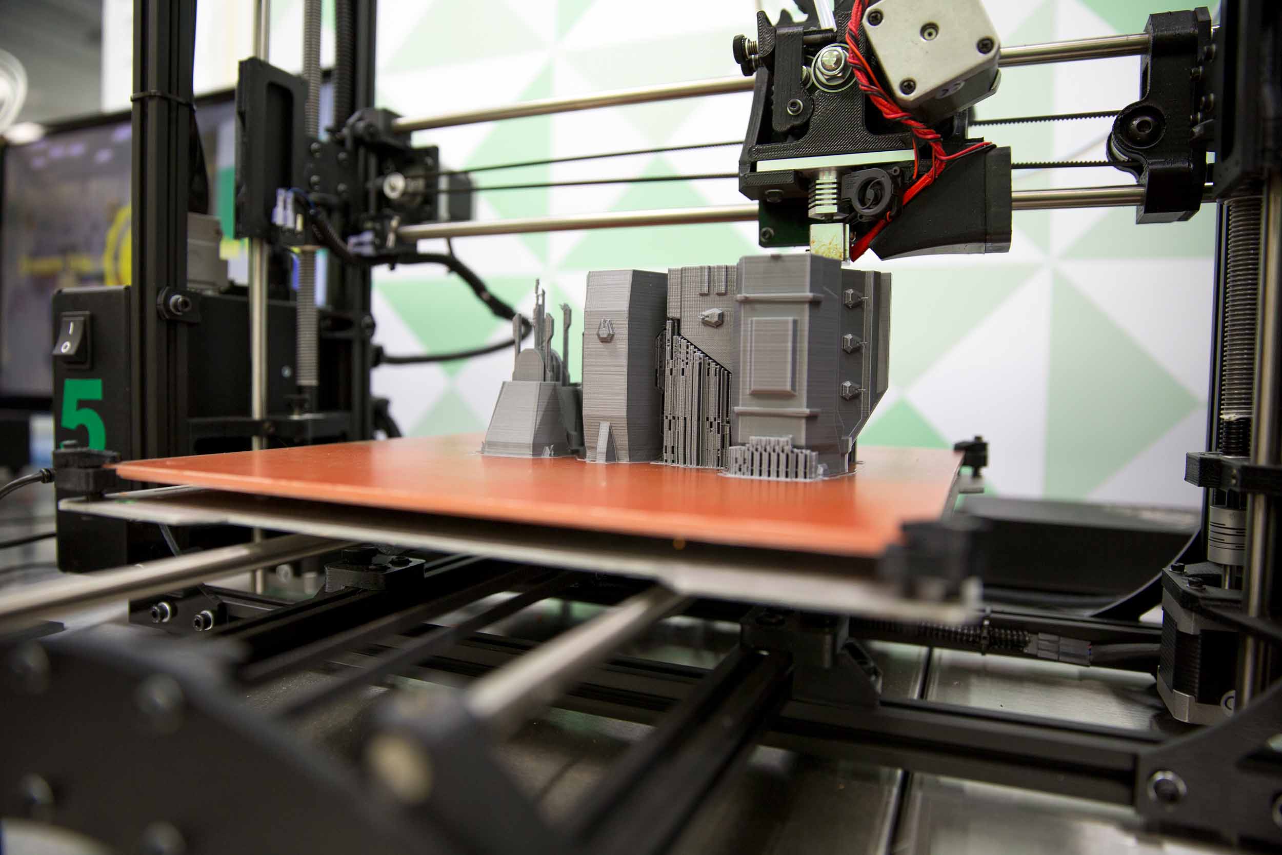 An additive manufacturing machine is shown in process of "printing" a grey object which looks a little like a model of a futuristic building